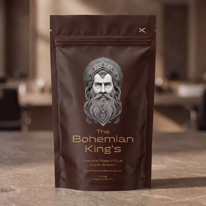 A stand-up pouch of beard dye standing on a stone-like surface with a blurred indoor background. The package is dark brown with a large silver-gray illustrated face and the text 'The Bohemian King's' and 'Natural Beard Dye Dark Brown'.