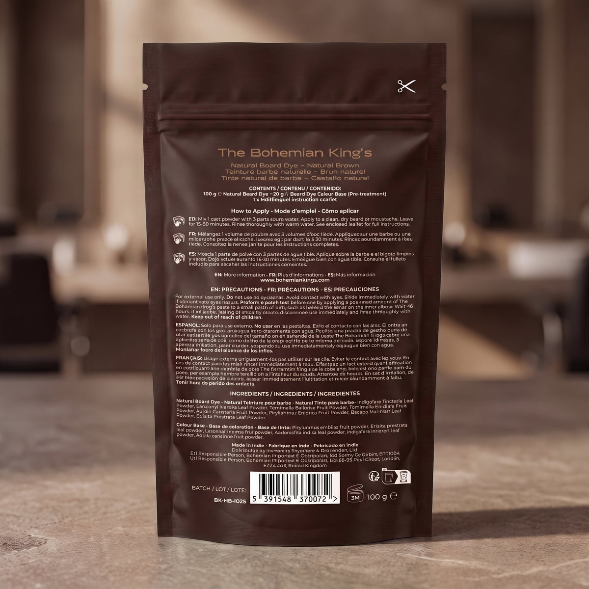 The back of a dark brown stand-up pouch for The Bohemian King's Natural Beard Oil, featuring product details, instructions, and a barcode, set against a blurred warm-toned indoor background.