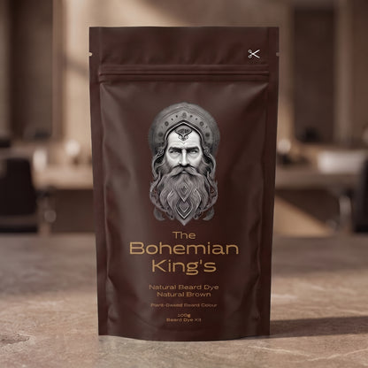 A stand-up pouch of beard dye standing on a stone-like surface with a blurred indoor background. The package is dark brown with a large silver-gray illustrated face and gold text reading 'The Bohemian King's' and 'Natural Beard Dye Natural Brown.