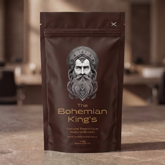 A stand-up pouch of beard dye standing on a stone-like surface with a blurred indoor background. The package is dark brown with a large silver-gray illustrated face and gold text reading 'The Bohemian King's' and 'Natural Beard Dye Natural Brown.
